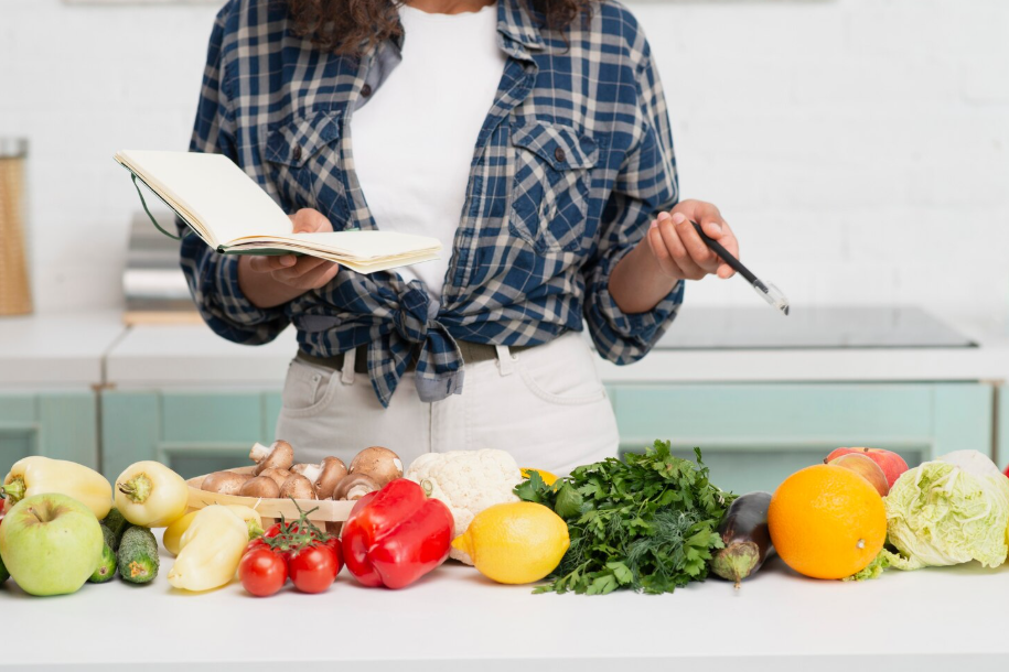 Person in holding a notebook and pen while planning budget meals, with fresh fruits and vegetables laid out.