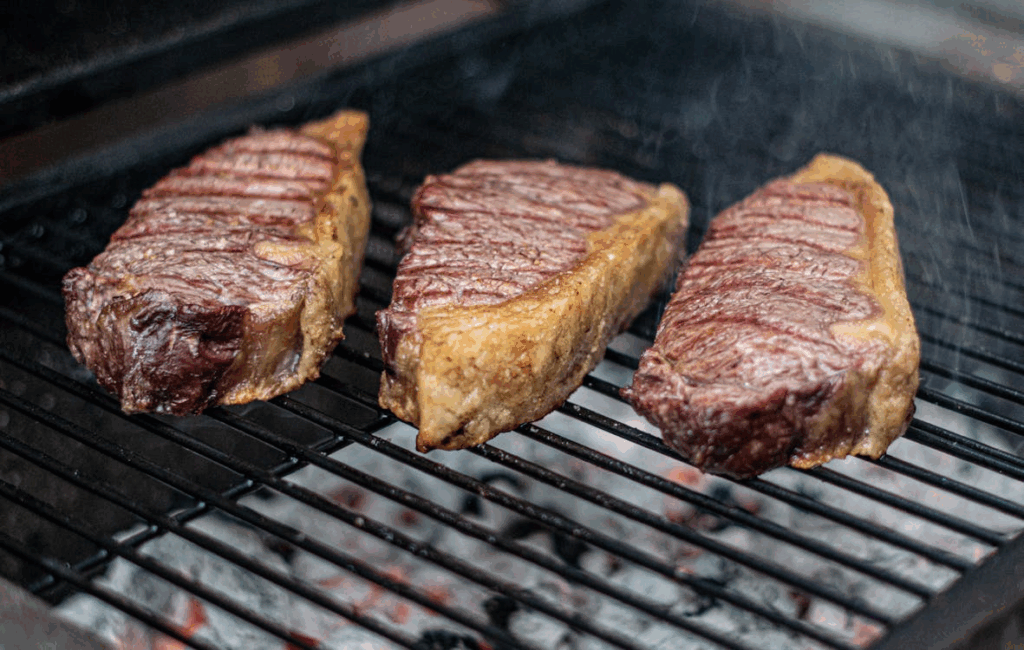 Three grilled steaks on a barbecue showing how to tell if meat is done by color and texture.
