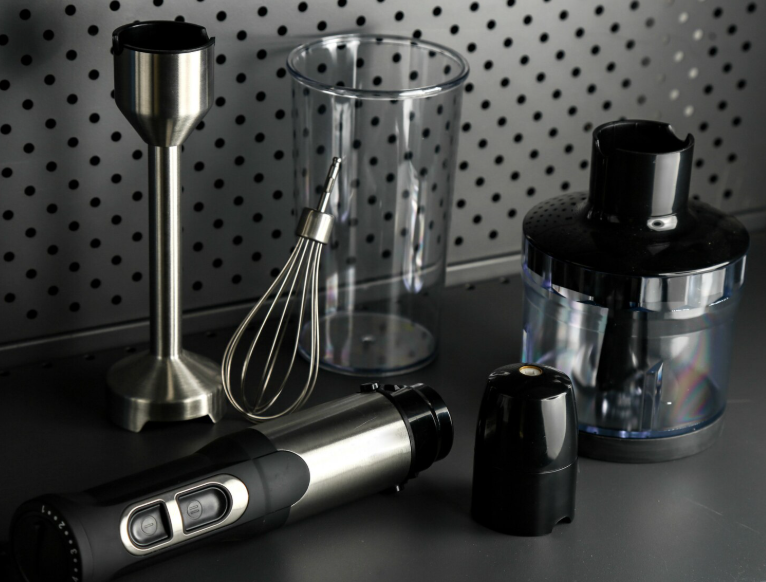 Immersion blender set with whisk, beaker, and chopper attachments on a modern kitchen counter.