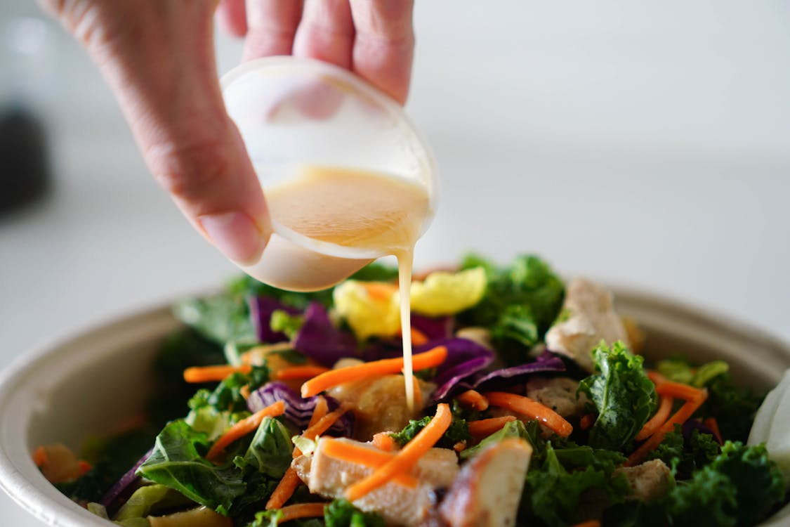 Close-up of a hand pouring creamy homemade salad dressing over a fresh green salad.