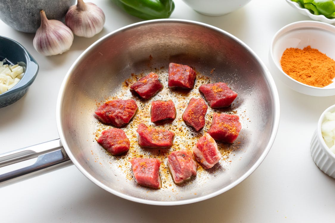 Overhead view of raw beef cubes sprinkled with seasoning in a stainless steel pan, ready for searing.
