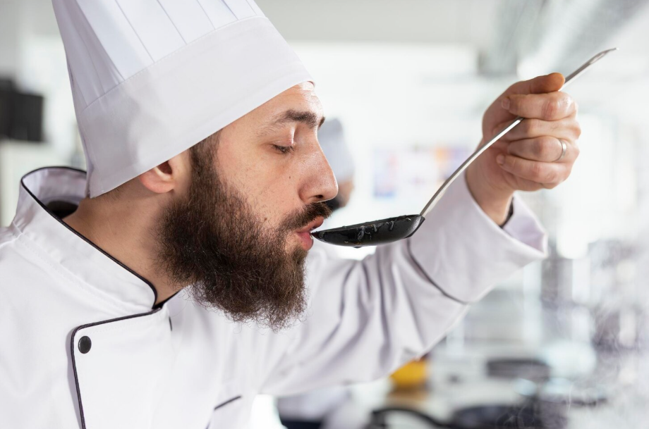 Chef in white uniform and hat tasting sauce from a spoon, checking flavor balance before serving.