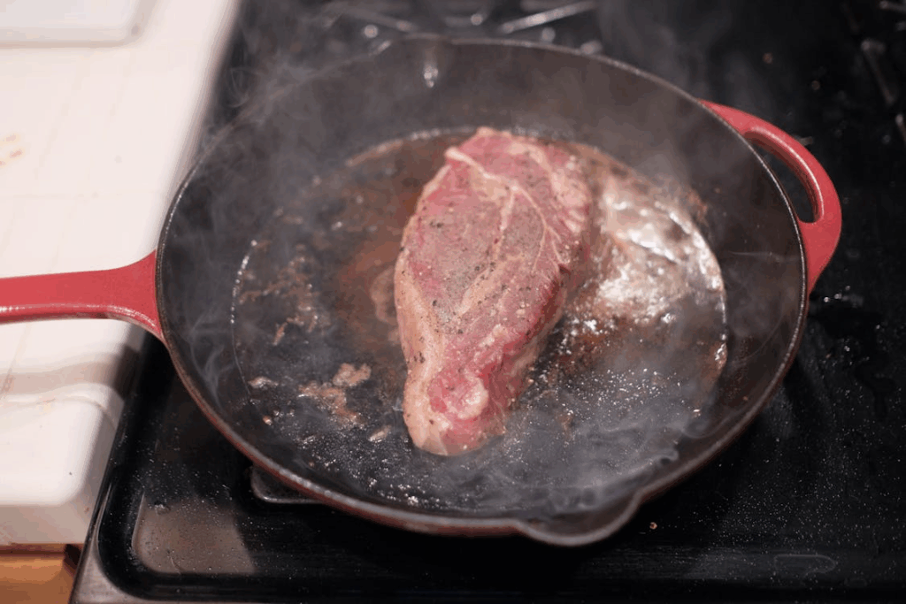 Thick steak sizzling in a hot cast iron pan with visible steam and browning crust.
