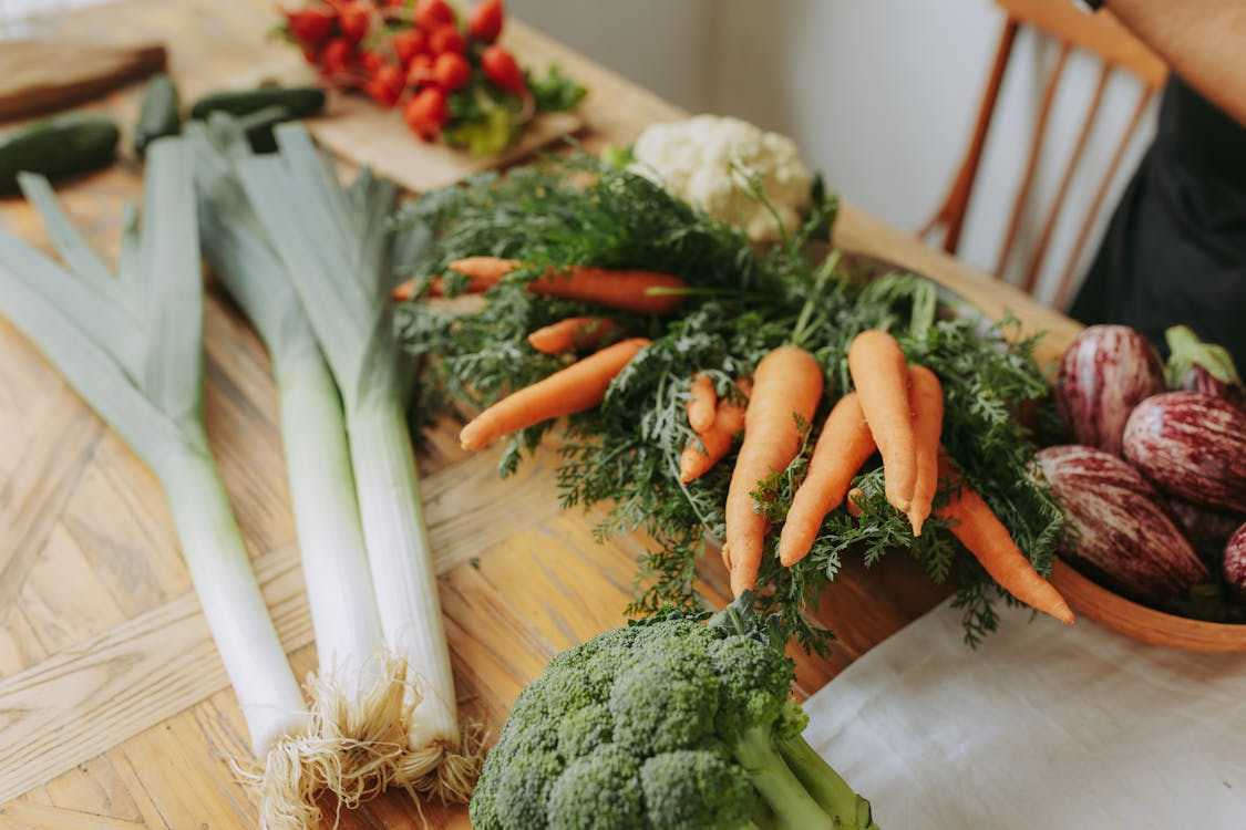 Assorted seasonal vegetables including leeks, carrots, broccoli, and eggplants arranged on a wooden table.