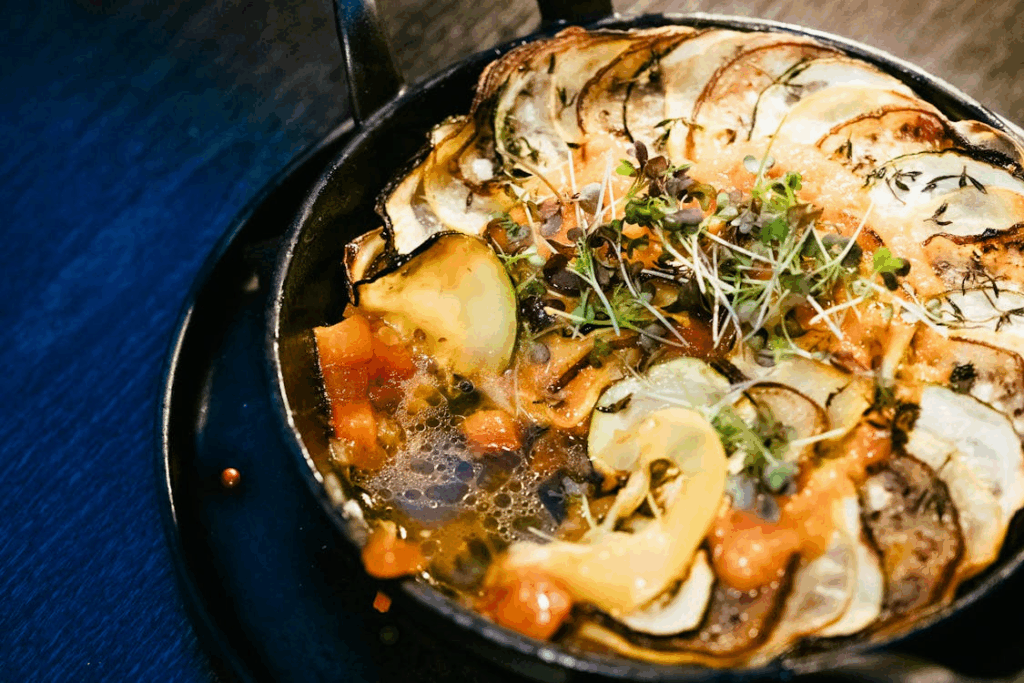 Old world recipes ratatouille-style vegetable bake in a cast-iron pan, layered potatoes and zucchini in tomato sauce.